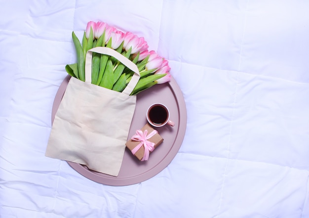 Vassoio con tazza di caffè, confezione regalo ed eco-bag con tulipani rosa sul lenzuolo bianco sul letto.