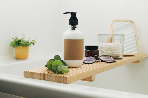 Vassoio in legno per vasca da bagno con flaconi cosmetici di bellezza e accessori in un moderno bagno scandinavo interno con piastrelle bianche come sfondo. Decorazioni per la casa con candele e piante verdi.