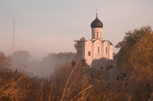 Vecchia chiesa ortodossa russa illuminata dal sole che sorge sopra la foresta autunnale in una leggera nebbia