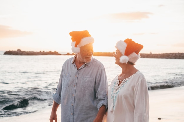 Vecchia coppia carina di persone mature che si divertono e si divertono insieme in spiaggia indossando cappelli di Natale nei giorni di vacanza Camminando sulla spiaggia con il tramonto sullo sfondo in invernoxA