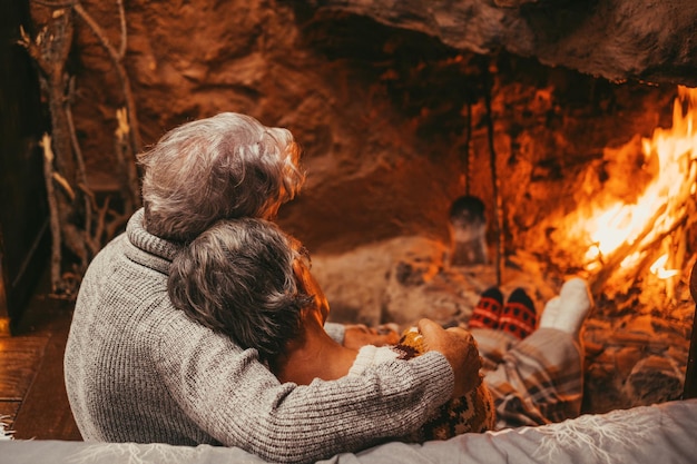 Vecchia coppia caucasica che trascorre il tempo libero insieme a casa sul retro dell'amorevole marito romantico che abbraccia la moglie e si rilassa guardando il caminetto acceso durante le vacanze invernali