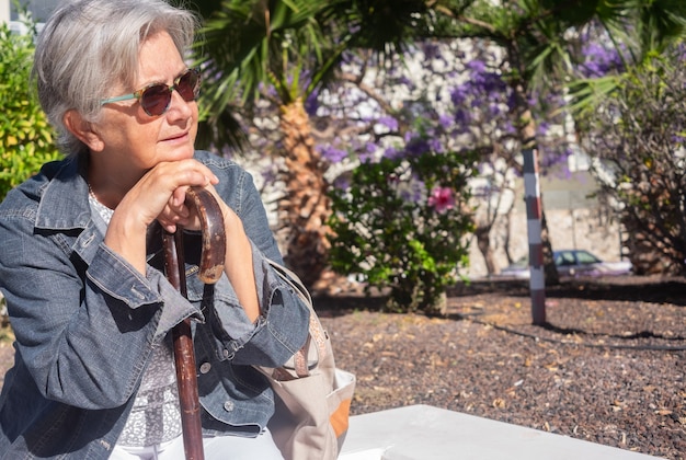 Vecchia donna anziana con le mani sopra il bastone seduto in un parco pubblico con espressione triste. Pensionato anziano con mal di schiena usando il bastone da passeggio. Albero e fiori sullo sfondo