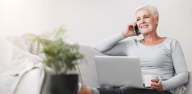 Vecchia donna attiva che tiene in mano un portatile e parla con lo smartphone