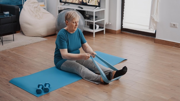 Vecchia donna che allunga braccia e gambe con fascia di resistenza, seduta su un tappetino da yoga. Adulto in pensione che tira la cintura elastica per fare esercizio fisico e sport. Allenamento donna con attrezzi fitness