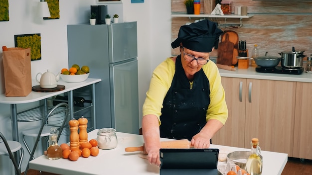 Vecchia donna con bonete che partecipa alla master class culinaria sul laptop. Chef in pensione seguendo i consigli del fornaio su tablet, imparando tutorial di cucina sui social media, usando il mattarello per formare l'impasto.