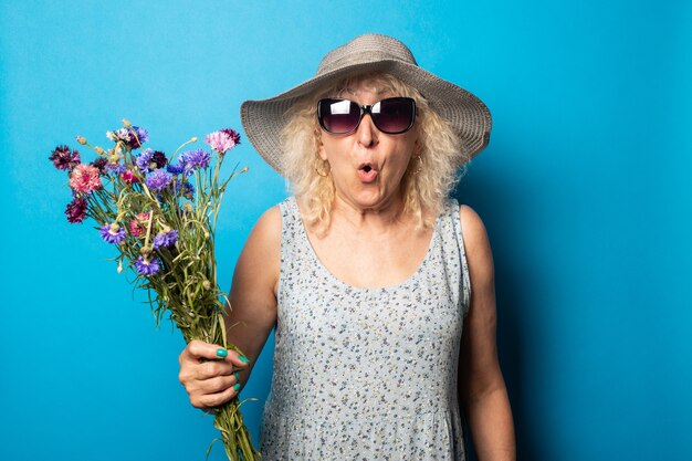 Vecchia donna con una faccia sorpresa in un cappello a tesa larga e un vestito che tiene un mazzo di fiori su una parete blu.