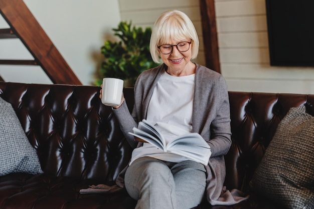 Vecchia donna in un libro di lettura casual che tiene tazza e sorride mentre è seduto in soggiorno sul divano