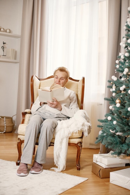 Vecchia donna seduta su una sedia il giorno di Natale. Nonna che legge un libro Donna che indossa abiti beige.