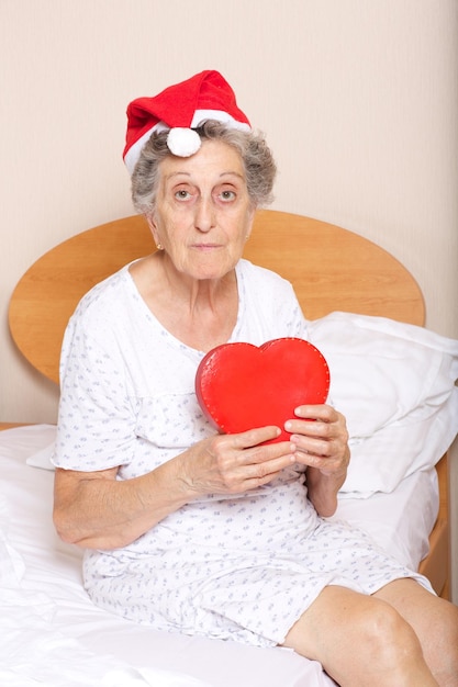 Vecchia donna tra i 70 e gli 80 anni vestita con cappello da Babbo Natale con cuore rosso tra le mani