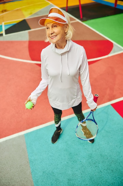 Vecchia, sana e allegra signora su una piazza colorata all'aperto e con in mano una racchetta da tennis con la palla