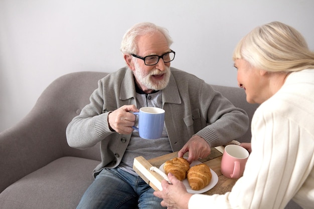 Vecchia signora con i capelli grigi che offre al marito bevande calde e croissant per il pasto mattutino. Nonno anziano con i capelli grigi che beve caffè portato da sua moglie in soggiorno.