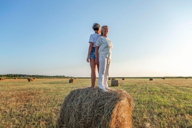 Vecchie e giovani donne sorridenti stanno su un rotolo di fieno in un campo al tramonto a raccogliere lavoro e libertà