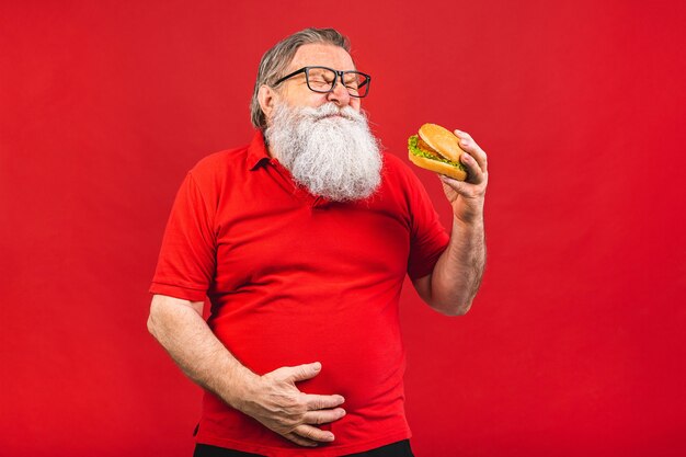 vecchio barbuto con gli occhiali che mangia un hamburger che tiene la sua pancia isolata su fondo rosso