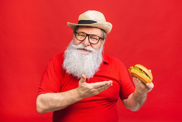 vecchio barbuto con gli occhiali in maglietta rossa che tiene un hamburger isolato su priorità bassa rossa