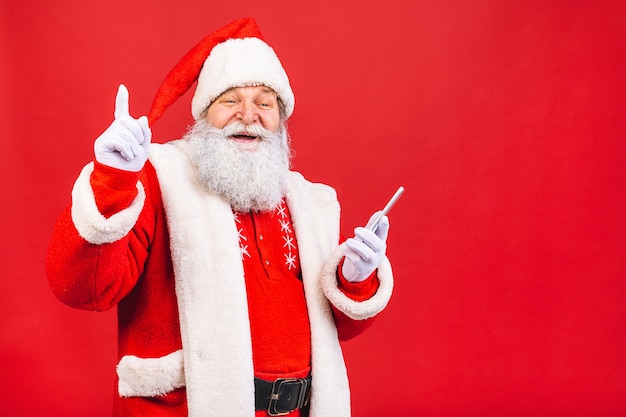 vecchio in costume di Babbo Natale che tiene su un telefono cellulare isolato su sfondo rosso