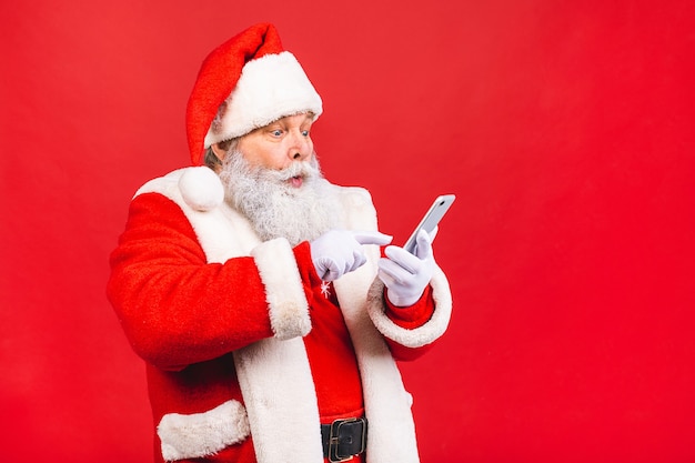 vecchio in costume di Babbo Natale che tiene su un telefono cellulare isolato sul rosso