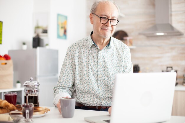 Vecchio in cucina che lavora da remoto. La vita quotidiana dell'uomo anziano in cucina durante la colazione utilizzando il computer portatile che tiene una tazza di caffè. Anziano pensionato che lavora da casa, telelavoro utilizzando il telecomando