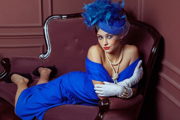 Vecchio stile retrò fotografia in stile Gran Bretagna. Bella giovane modella caucasica in abito blu e trucco moda e cappello in posa seduto sul divano artificiale e guardando in lontananza.
