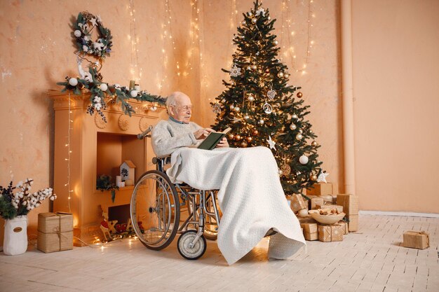 Vecchio su una sedia a rotelle seduto vicino all'albero di Natale e leggendo un libro