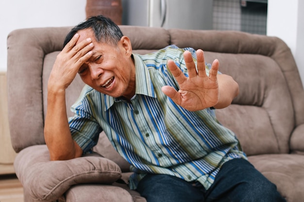 Vecchio uomo anziano seduto sul divano nel soggiorno che tocca la fronte e soffre di forti mal di testa