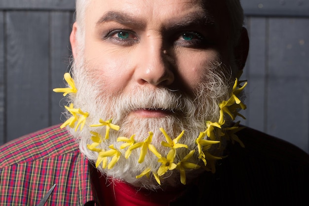 Vecchio uomo barbuto con fiori gialli in lunga barba grigia sul viso sorridente in camicia a scacchi rossa su sfondo di legno