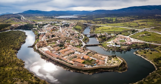 Veduta aerea di Buitrago del Lozoya, un villaggio storico ben conservato vicino a Madrid, circondato dal fiume Lozoya, con la catena montuosa del Guadarrama e il bacino idrico sullo sfondo.