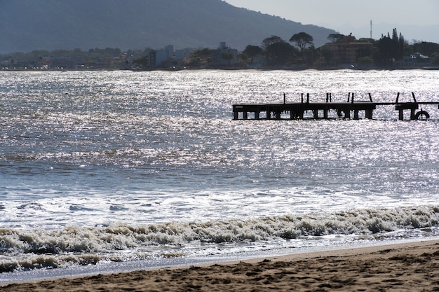 Veduta della spiaggia di Rio das Ostras con la riunione del fiume a Rio de Janeiro. Giornata di sole, cielo blu. Sabbia gialla e alcune rocce. Ponte in legno da attraversare. Barche e pesca sul molo.