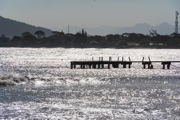 Veduta della spiaggia di Rio das Ostras con la riunione del fiume a Rio de Janeiro. Giornata di sole, cielo blu. Sabbia gialla e alcune rocce. Ponte in legno da attraversare. Barche e pesca sul molo.