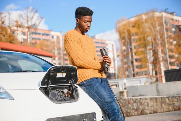 Veicolo urbano, elettrico, concetto di eco. Giovane uomo dalla pelle nera, in attesa che la sua auto elettrica si ricarichi