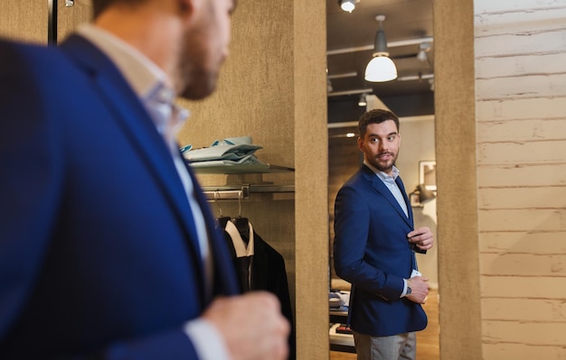 vendita, shopping, moda, stile e concetto di persone - giovane elegante che sceglie e prova la giacca e cerca di specchiarsi nel centro commerciale o nel negozio di abbigliamento