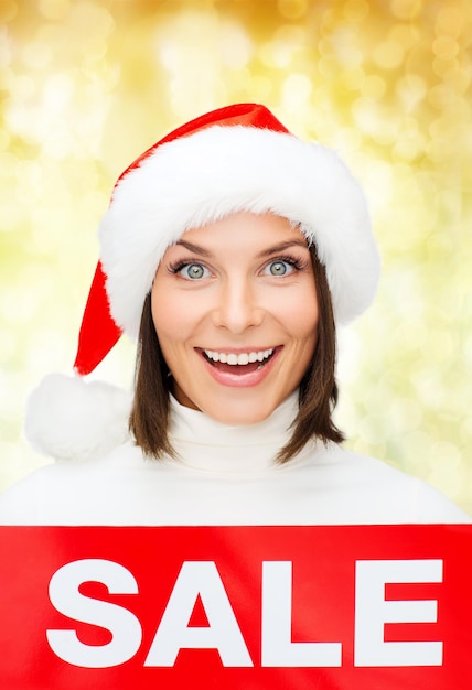 vendita, shopping, natale, vacanze e concetto di persone - donna sorridente in cappello santa helper con segno di vendita rosso su sfondo di luci gialle