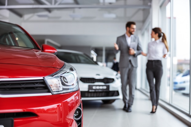 Venditore di auto maschio in vestito che cammina intorno al salone dell'auto con la donna che vuole comprare un'auto e parlando delle specifiche delle auto