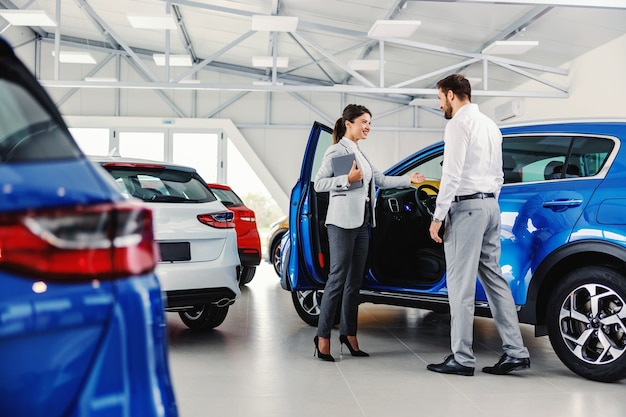 Venditore femminile amichevole e sorridente che mostra un'auto nuova di zecca a un cliente mentre si trova nel salone dell'auto