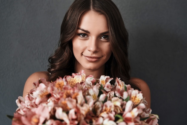 Vera bellezza. Attraente giovane donna con bouquet di fiori che guarda l'obbiettivo e sorride mentre sta in piedi su sfondo grigio gray