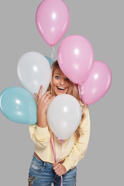 Vera felicità. Attraente giovane donna sorridente che si nasconde tra palloncini e guarda la telecamera mentre si trova in piedi su sfondo grigio gray