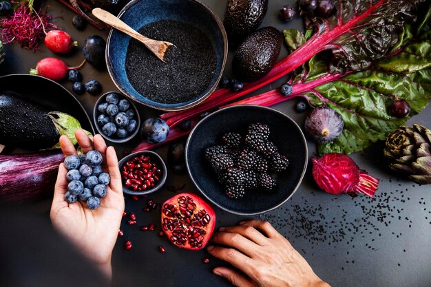 Verdure e frutta crude viola su sfondo nero e una mano che tiene i mirtilli