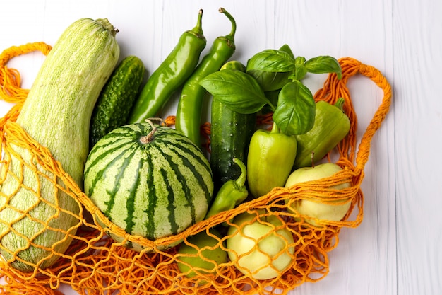 Verdure e frutta verdi in borsa shopping mesh riutilizzabile arancione su superficie bianca: zucchine, cetrioli, peperone, peperoncino, mele, pera, anguria e basilico, orientamento orizzontale