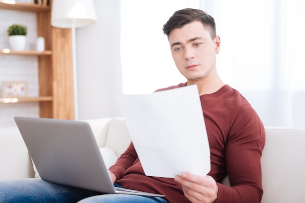 Verifica sempre. Studente maschio piacevole sicuro che tiene carta durante l'utilizzo di laptop