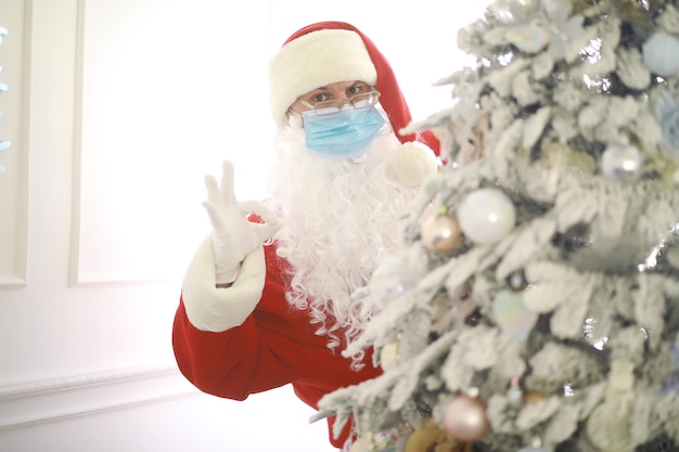 Vero Babbo Natale e albero di Natale su uno sfondo, con indosso una maschera protettiva, occhiali e cappello. Natale con distanza sociale. Covid-19