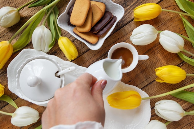 Versare a mano il latte nel caffè vicino ai tulipani bianchi e gialli sul tavolo in legno