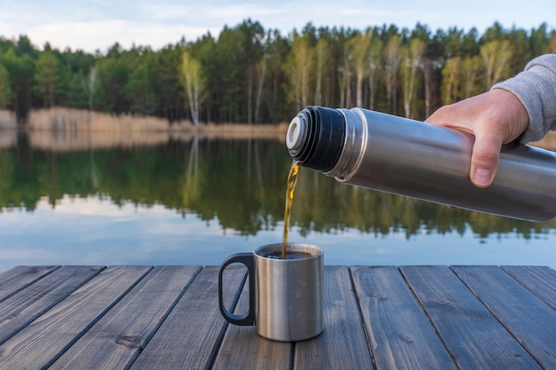 Versare il tè caldo in una tazza da un thermos al mattino vicino al lago e alla foresta in primavera, primo piano. Natura e concetto di viaggio