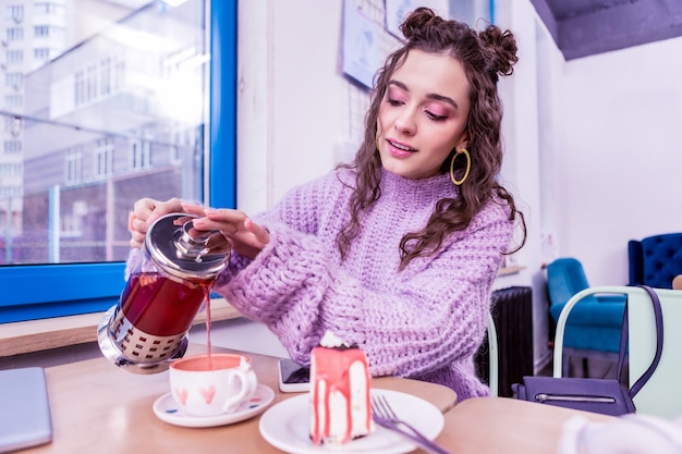 Versare il tè caldo. Ragazza dai capelli scuri concentrata che aggiunge altro tè nella tazza decorata con cuori acquerellati