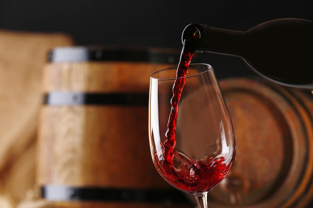 Versare il vino rosso dalla bottiglia nel bicchiere con botti di vino in legno sullo sfondo