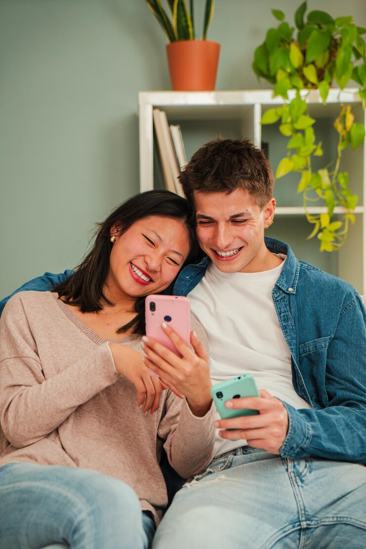 Vertical multiracial married couple using a cellphone to search apps sitting on living room sofa