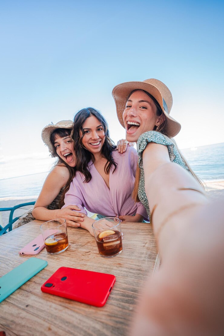 Vertical three girls using a mobile phone to take a selfie sitting at a beach bar terrace a group of