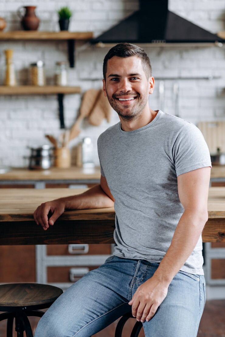Photo vertical view of the handsome man sitting on the chair leaned on table, smiling and looking at the camera. image at the cozy kitchen