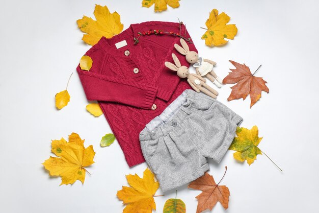 Vestiti alla moda del bambino di autunno con foglie e giocattoli sulla superficie bianca