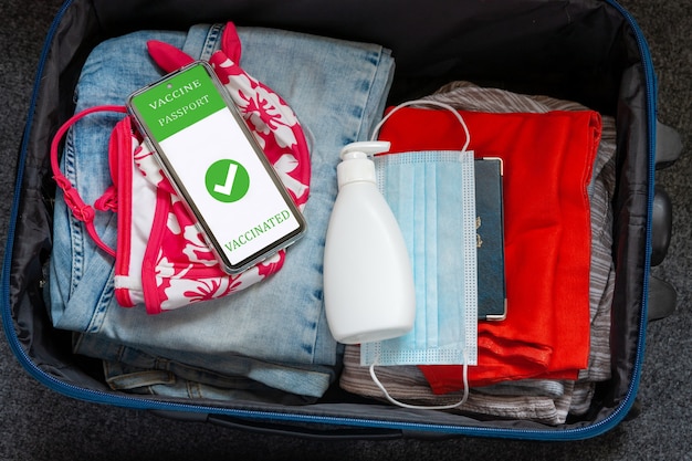 Vestiti per i viaggi di vacanza nella borsa da viaggio, maschera protettiva per il viso, liquido disinfettante nell'erogatore e identificazione digitale del passaporto del vaccino nel telefono cellulare. Nuovo concetto di viaggio normale