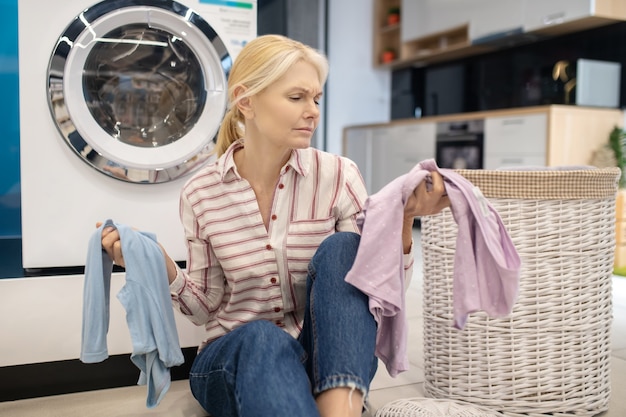 Vestiti puliti. Casalinga bionda in camicia a righe che si siede vicino alla lavatrice e che tiene i vestiti