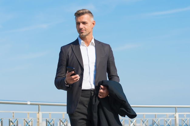 Vestito formale dell'uomo d'affari con lo smartphone. Concetto di vita moderna. Capo sulla strada per l'ufficio. Usa il telefono mentre cammini. Stile urbano. Giornata lavorativa regolare. Fondo del cielo blu dell'uomo carismatico. Telefonata.